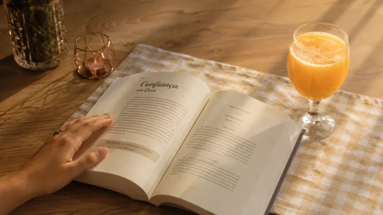 Mão acompanhando leitura de devocional aberto sobre toalha xadrez em mesa de madeira clara, com suco de laranja ao lado